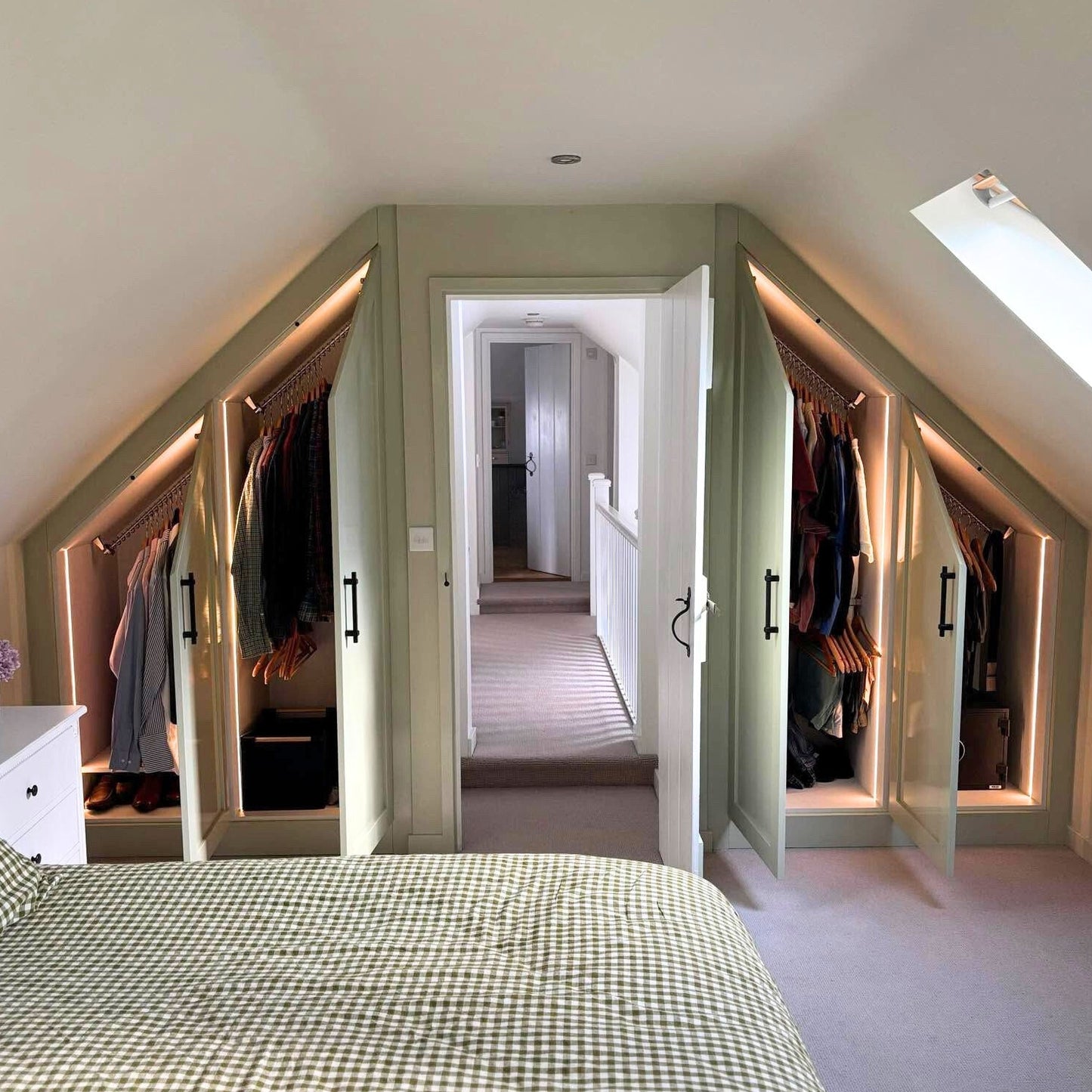 Green sloped ceiling wardrobes, to either side of a doorway in a loft bedroom, top fitted with angled clothes rails with hangers and clothes with shoes below