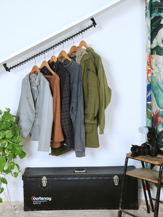 A black clothes rail fitted to an angled ceiling with clothes hanging from it on hangers, storage boxes, plant curtains and stools