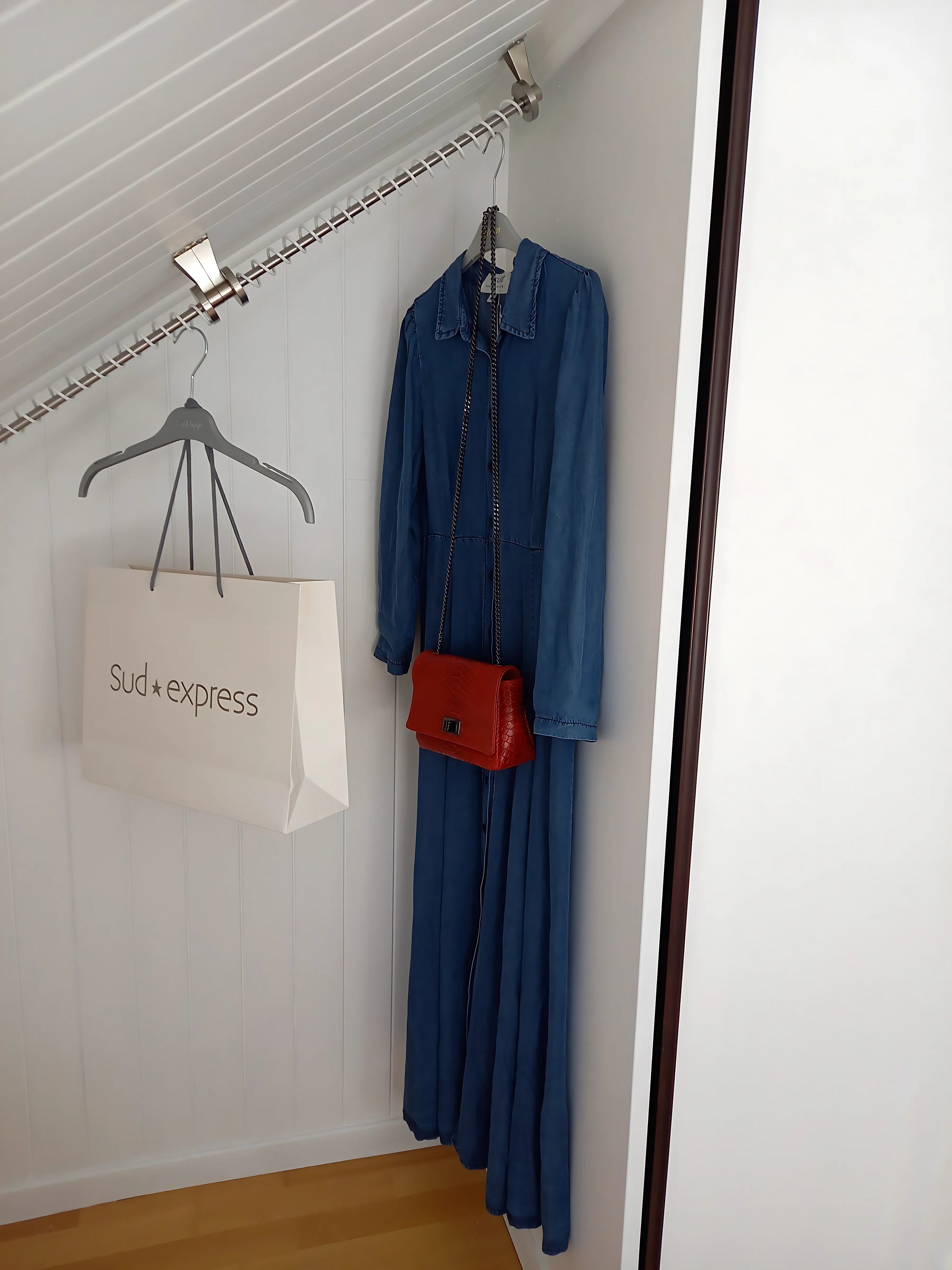 Blue coat, red handbag, and beige bag with 'sud express' logo hanging on a top fixed sloping clothes rail