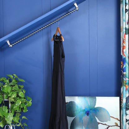 Black dress hanging on a Zebedee hanging rail against an angled blue wall in an attic room with a plant and artwork in the foreground.