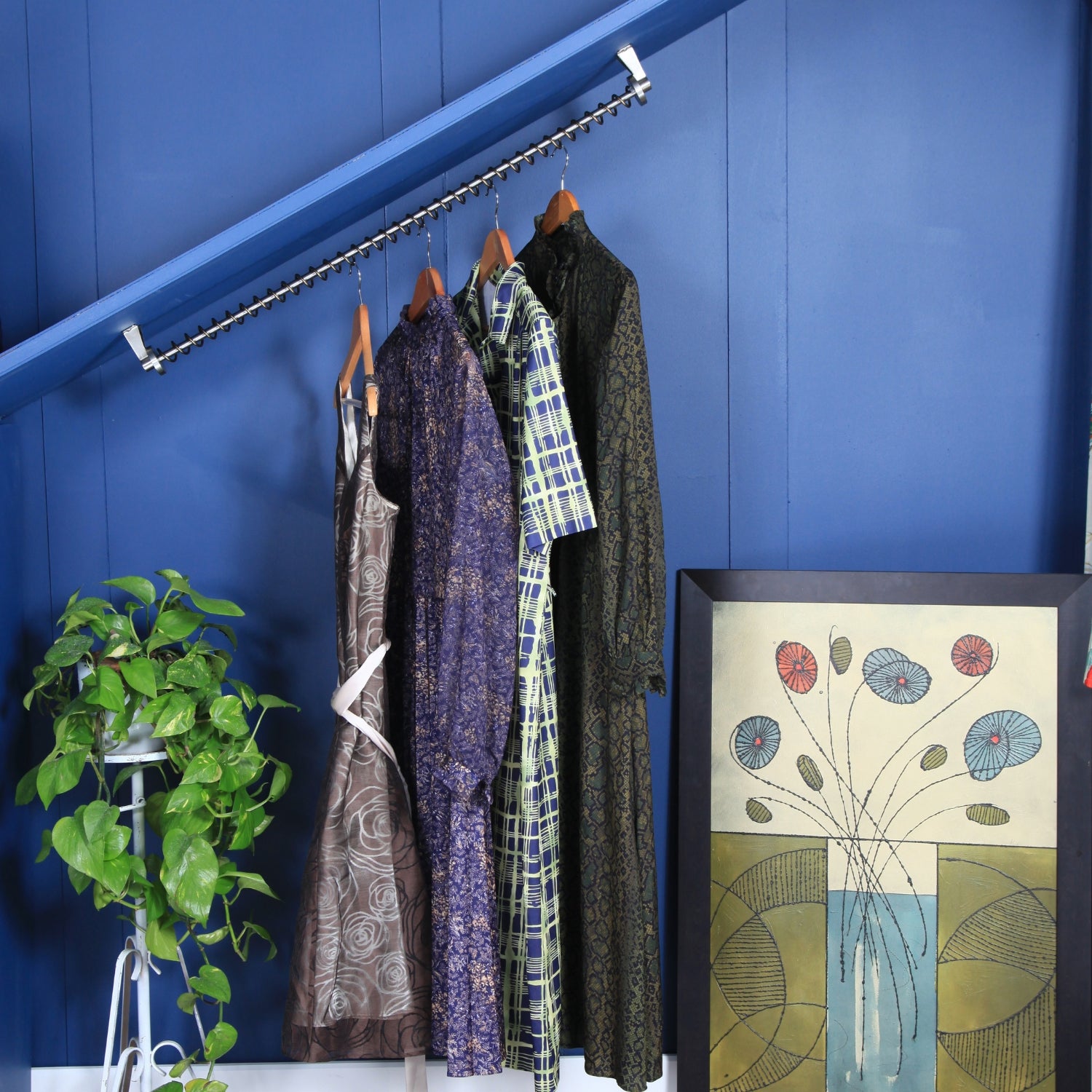 Sloping clothing hanging rail  top mounted on an angled ceiling against a blue wall with a plant and artwork.