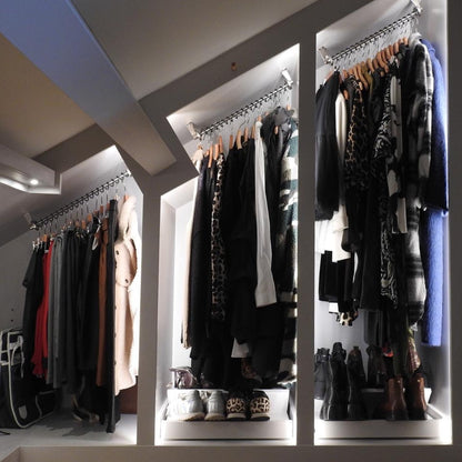 Illuminated internal view of a sloped ceiling fitted wardrobe with angled wardrobe rails, clothes on hangers and shoes on a shelf.