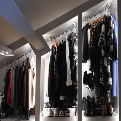 An open sloped ceiling wardrobe walk in dressing room with top fixed angled clothes rails, clothes hanging full length with shoe racks beneath
