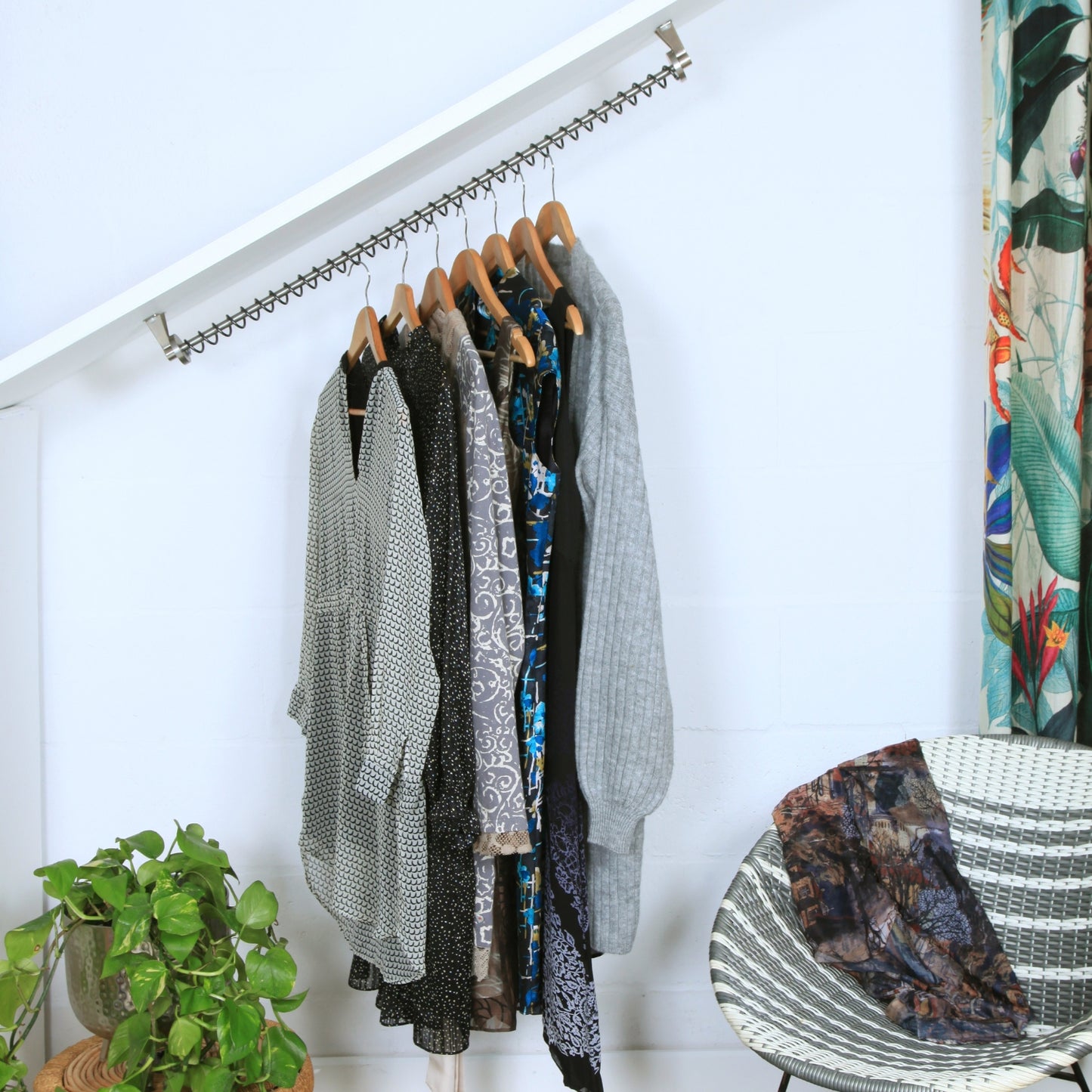 Angled clothing rack with various garments against a white wall, next to a chair and plant.